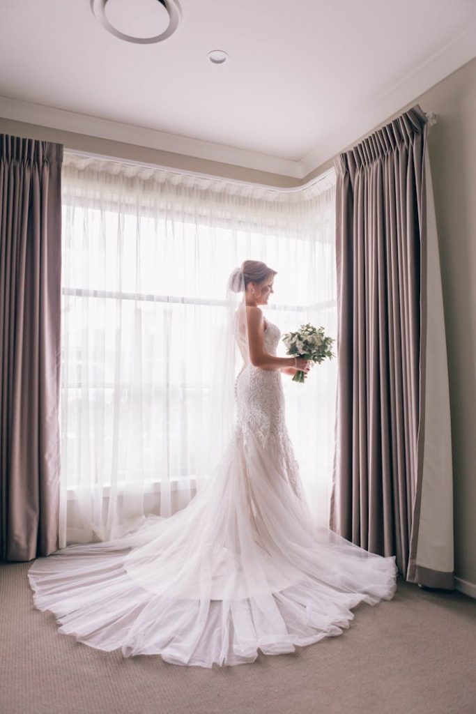 Beautiful bride in white wedding gown posing indoors by a window.